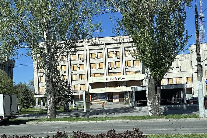 Ein früheres Kino namens „кіно Байда“ steht inmitten einer Baumallee. Die Fenster sind fast vollständig mit Holzplatten verbarrikadiert, während am Eingang alte Filmplakate sichtbar sind. Der Ort wirkt verlassen, einzelne Menschen bewegen sich im Hintergrund.