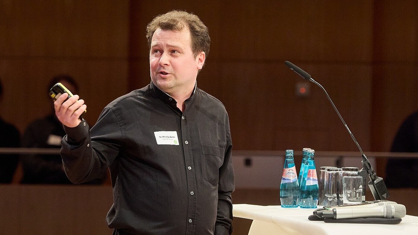 Herr Belov hält eine Fernbedienung in der Hand und deutet hinter sich auf den Bildschirm.