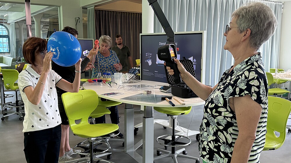 Zwei Lehrerinnen testen Versuche im School Lab. Eine hält einen blauen Luftballon vor ihr Gesicht, die andere hat ein digitales Messgerät in der Hand.