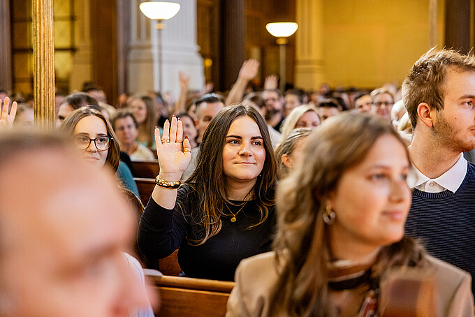 Eine junge Frau sitzt zwischen weiteren Gästen in einer Kirchenbank und hebt die Hand.
