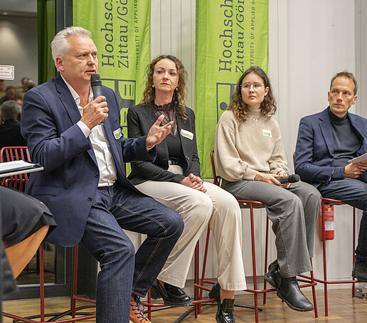 Im Panel sitzen vier Gäste auf Hochstühlen nebeneinander, ein Gast spricht in ein Mikrofon.