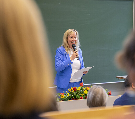 Die Prorektorin im blauen Anzug spricht in ein Mikrofon vor einer grünen Tafel im Hörsaal.