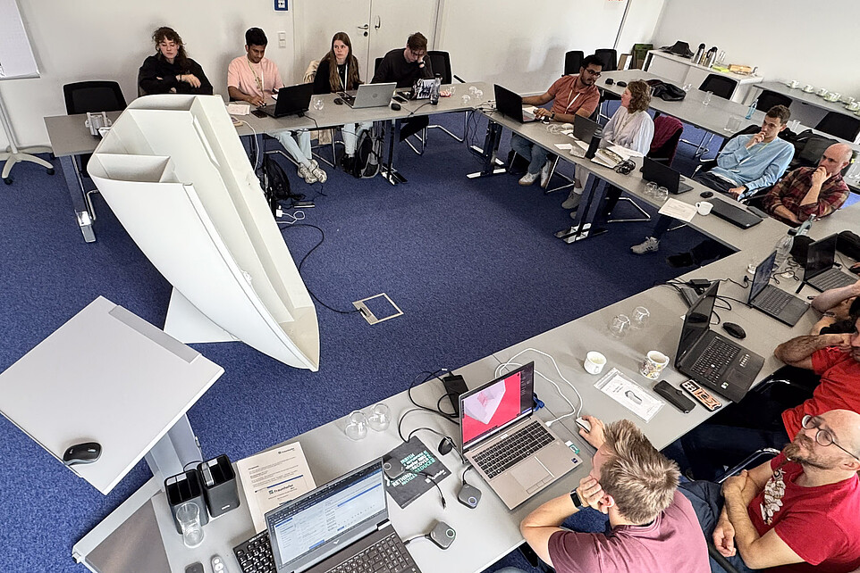 Teilnehmende sitzen mit Laptops an Tischen rund um ein weißes Boot im Seminarraum.