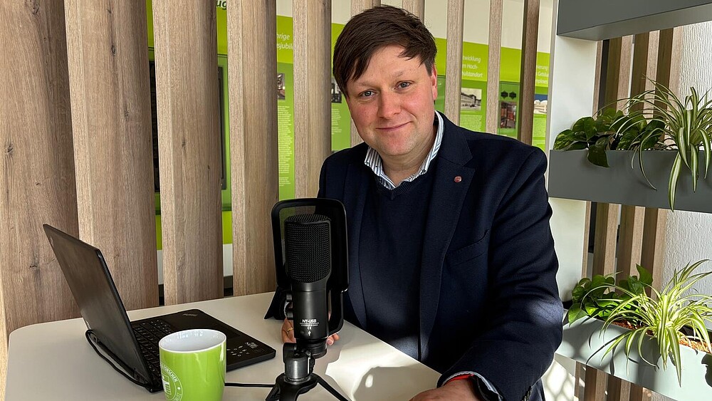 Daniel Winkler sitzt vor einem Mikrofon und Laptop an einem Stehtisch und lächelt in die Kamera.