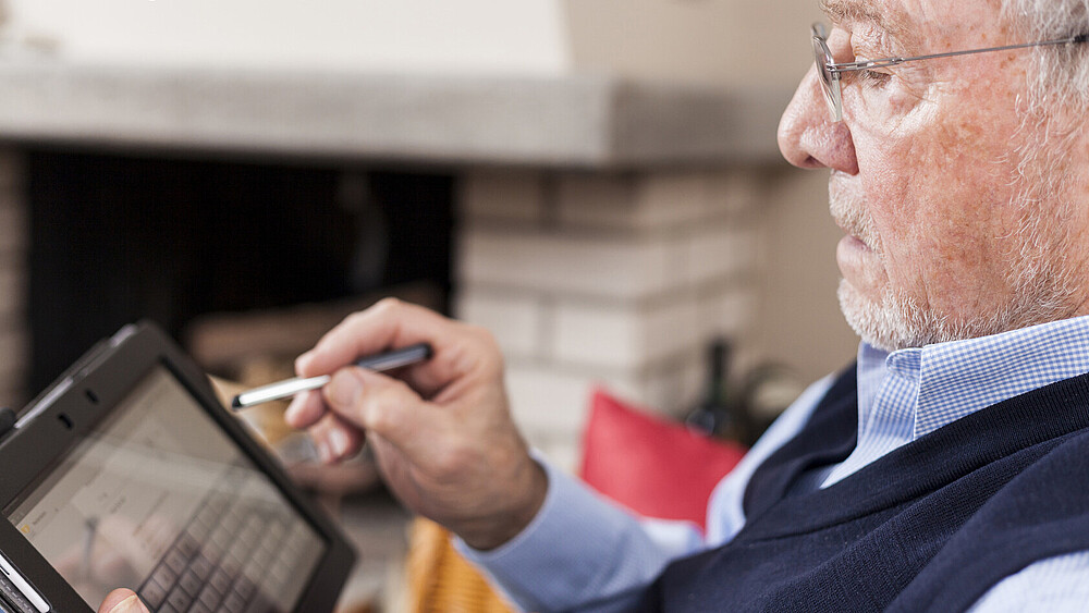 Ein älterer Herr mit Brille hält ein Tablet in einer Hand und einen Stift in der anderen Hand, mit dem er das Display bedient.